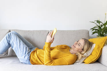 Woman using smart phone while lying on sofa at home - WPEF04984