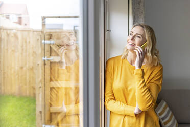 Lächelnde Frau, die in der Nähe der Wohnungstür mit einem Smartphone telefoniert - WPEF04990