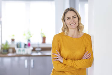 Smiling woman with arms crossed leaning on wall at home - WPEF04999