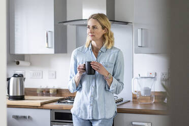 Woman with tea cup looking away while standing in kitchen at home - WPEF05002