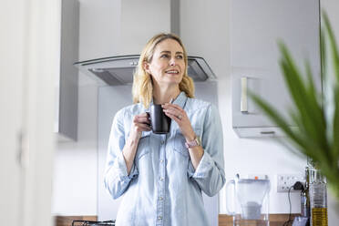 Blonde Frau hält Tasse, während sie in der Küche zu Hause wegschaut - WPEF05003