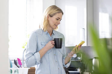 Woman with mug using mobile phone in kitchen at home - WPEF05007
