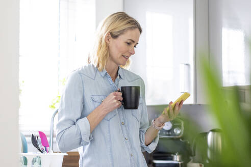 Frau mit Becher benutzt Handy in der Küche zu Hause - WPEF05007