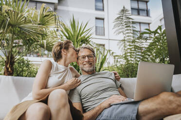 Frau küsst Mann, der mit Laptop auf einem Sofa auf der Terrasse sitzt - MFF08211