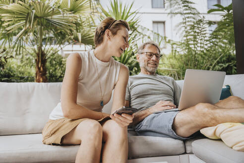 Frau im Gespräch mit Mann, der auf der Terrasse am Laptop arbeitet - MFF08213