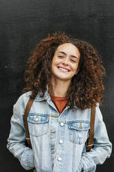 Smiling young curly haired woman standing with hands in pockets - JCMF02022