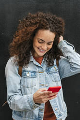Smiling young woman text messaging through smart phone while standing with hand in hair - JCMF02024