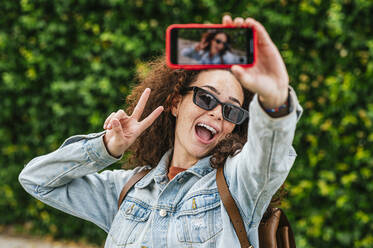 Junge Frau, die ein Selfie mit ihrem Smartphone macht, während sie das Friedenszeichen mit offenem Mund zeigt - JCMF02040