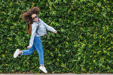 Excited young woman wearing sunglasses jumping in front of green ivy plants - JCMF02041