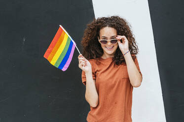 Lächelnde junge Frau mit Sonnenbrille, die eine Regenbogenflagge vor einer Wand hält - JCMF02046