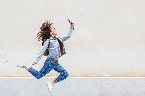 Aufgeregte Frau, die ein Selfie mit ihrem Smartphone macht, während sie vor eine Wand springt - JCMF02052
