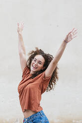 Fröhliche junge Frau tanzt mit erhobenen Armen vor einer Wand - JCMF02056