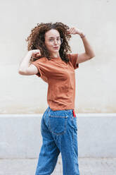 Selbstbewusste junge Frau, die vor einer Wand ihre Muskeln anspannt - JCMF02059