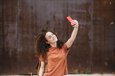 Smiling young woman taking selfie from mobile phone in front of wall - JCMF02060