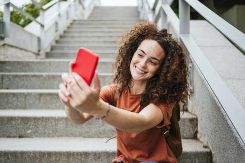 Schöne lächelnde Frau mit lockigem Haar unter selfie durch Smartphone sitzen auf Stufen - JCMF02062