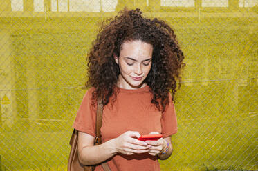 Junge Frau mit lockigem Haar, die mit ihrem Smartphone vor einem gelben Zaun Textnachrichten verschickt - JCMF02064