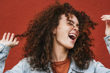 Cheerful young woman with curly hair singing in front of red wall - JCMF02077