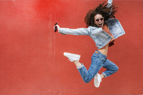 Aufgeregte junge Frau mit Sonnenbrille springt vor einer roten Wand - JCMF02083