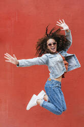 Glückliche Frau mit Sonnenbrille, die vor einer roten Wand springt - JCMF02084