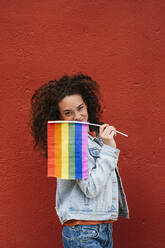 Junge Frau hält Regenbogenflagge vor roter Wand - JCMF02093