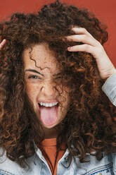 Young curly haired woman with hands in hair sticking out tongue - JCMF02098