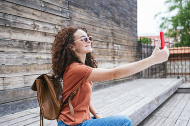 Smiling young woman wearing backpack taking selfie through smart phone - JCMF02105