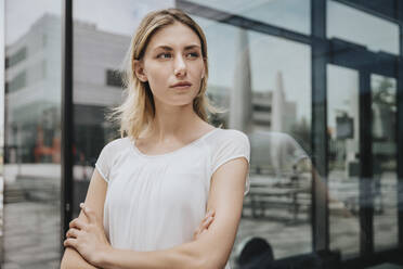 Serious woman with arms crossed looking away in front of glass wall - MFF08217