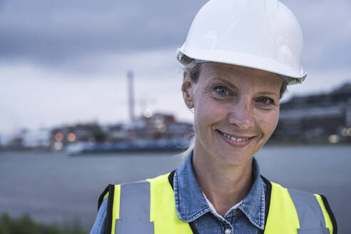 Smiling female professional wearing hardhat at sunset - UUF23779