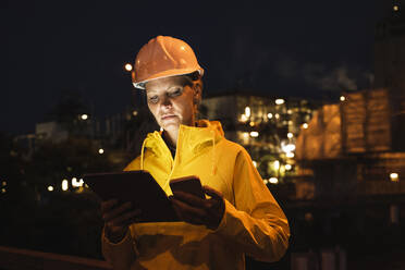 Female professional in yellow hardhat and raincoat using wireless technologies at night - UUF23812