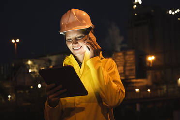 Smiling female professional talking on smart phone while holding digital tablet at night - UUF23813