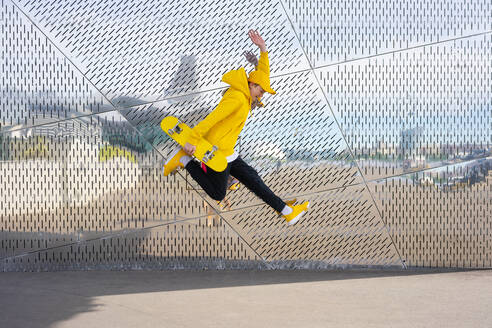 Boy jumping while holding skateboard by metal wall - VPIF04215