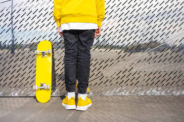 Male skater wearing yellow shoes standing by skateboard in front of metallic wall - VPIF04222