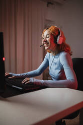 Seitenansicht einer begeisterten Spielerin mit Kopfhörern, die ein Videospiel spielt, während sie zu Hause am Tisch sitzt - ADSF25238