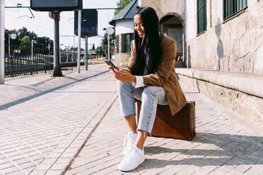 Cheerful Asian traveling female sitting on vintage suitcase on platform at railway station and browsing mobile phone while waiting for train - ADSF25241