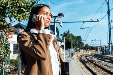 From below side view of content ethnic female traveler standing on platform at railroad station and talking on smartphone - ADSF25242