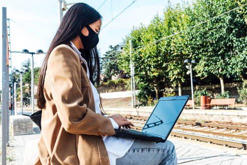 Seitenansicht einer asiatischen Frau mit Maske, die auf einer Bank am Bahnhof sitzt und am Laptop arbeitet, während sie auf den Zug wartet - ADSF25243