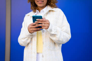 Cropped unrecognizable delighted African American female using smartphone while standing on blue background - ADSF25245