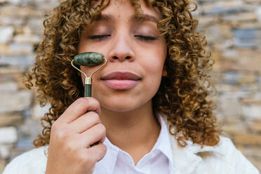 Dreamy African American female using jade roller and doing facial massage while standing with closed eyes in park - ADSF25251