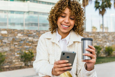 Positiv eingestellte Afroamerikanerin mit Kaffee zum Mitnehmen, die auf einer Straße in der Stadt über ihr Smartphone Nachrichten in sozialen Medien verschickt - ADSF25253