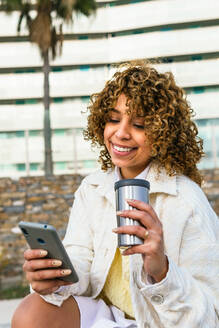 Positive African American female with takeaway coffee messaging on social media via smartphone in street of city - ADSF25254