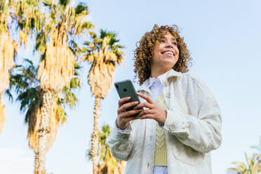 Low angle of cheerful African American female browsing mobile phone while standing in exotic park with palm trees at sunset and looking away - ADSF25256