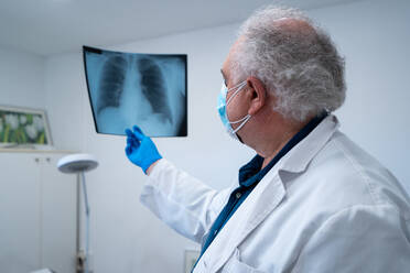 Seitenansicht eines älteren männlichen Arztes mit Maske und Handschuhen, der ein Röntgenbild der Lunge eines Patienten mit Coronavirus im Krankenhaus untersucht - ADSF25281