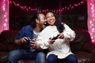 Aufgeregtes ethnisches Paar in Freizeitkleidung mit Freude Pads spielen Videospiel zusammen beim Sitzen auf Ledercouch zu Hause - ADSF25318