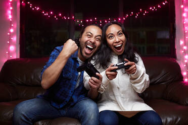 Aufgeregtes ethnisches Paar in Freizeitkleidung mit Freude Pads spielen Videospiel zusammen beim Sitzen auf Ledercouch zu Hause - ADSF25319