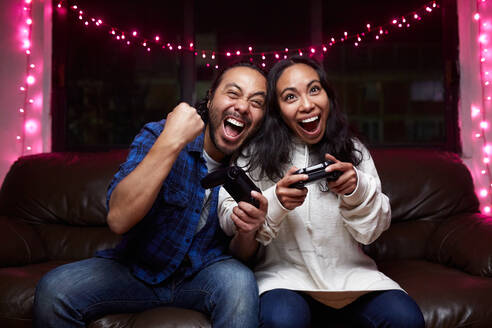 Excited ethnic couple in casual wear with joy pads playing video game together while sitting on leather couch at home - ADSF25319