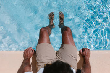 Top view of unrecognizable male freelancer sitting at poolside with legs in water during telework in summer - ADSF25320