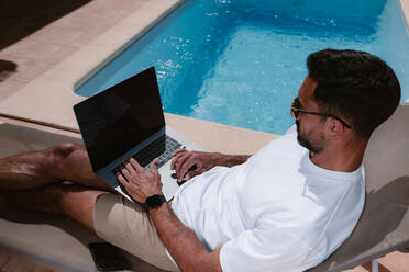 Männlicher Freiberufler liegt auf einer Liege am Pool und surft im Internet auf einem Laptop während der Telearbeit im Sommer an einem sonnigen Tag - ADSF25322