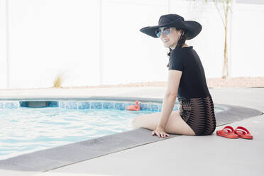 Lächelnde Frau mit Sonnenbrille, die wegschaut, während sie im Urlaub am Pool sitzt - ACTF00112