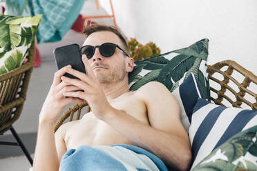 Mann mit Handtuch, der ein Mobiltelefon benutzt, während er sich auf dem Sofa im Innenhof entspannt - ACTF00115