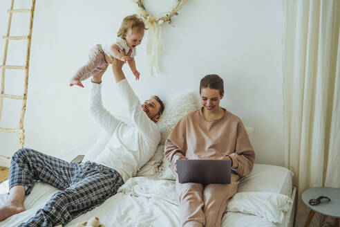 Smiling woman using laptop by playful man lifting daughter on bed at home - VPIF04293
